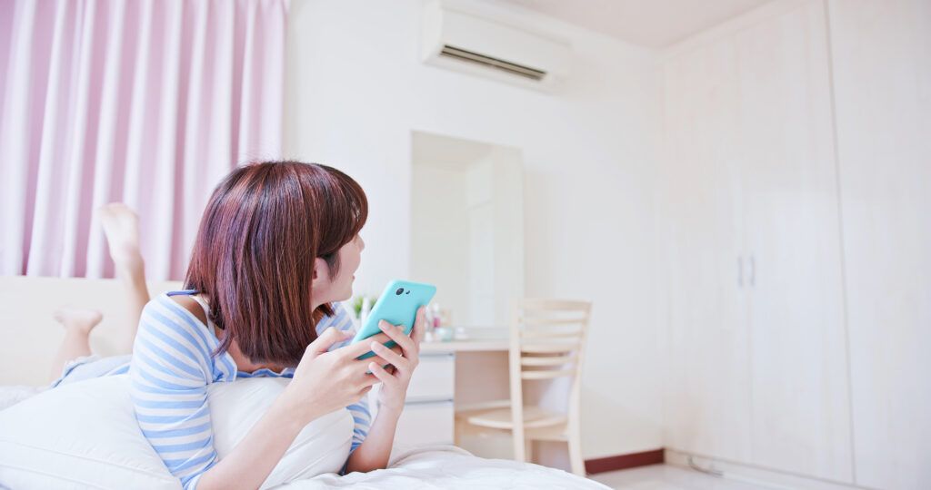 Mujer joven con smartphone descansando en un dormitorio luminoso con cortinas rosas y ambiente confortable.