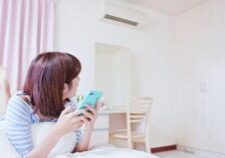 Mujer joven con smartphone descansando en un dormitorio luminoso con cortinas rosas y ambiente confortable.